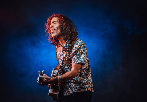 Hispanik müzisyen, mavi ve kırmızı ışıkla aydınlatılmış bir karanlıkta duygusal olarak şarkı söylüyor ve gitar çalıyor. - Fotoğraf, Görsel
