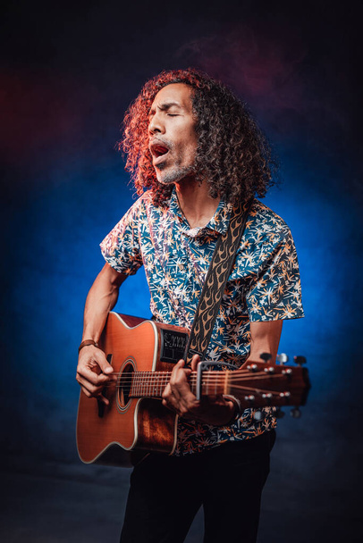 Músico hispano emocionalmente cantando y tocando la guitarra en una oscuridad iluminada por la luz azul y roja
 - Foto, Imagen
