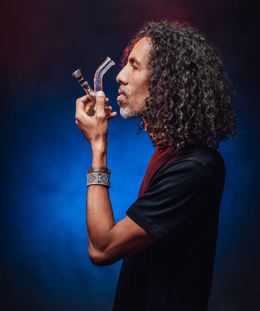 Fumo maschile ispanico di mezza età da bong su uno sfondo scuro illuminato luce blu - Foto, immagini
