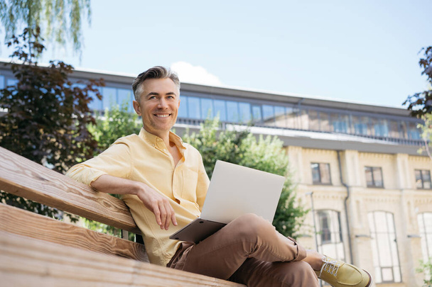 Komea kypsä liikemies käyttää kannettavaa tietokonetta, suunnittelee käynnistystä, istuu ulkona. Hymyilevä freelancer puistossa istumassa penkillä. Onnistunut liiketoimintakonsepti - Valokuva, kuva