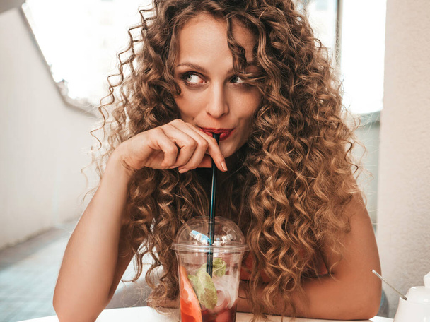 Young beautiful smiling hipster woman with afro curls hairstyle. Woman in summer trendy clothes sitting in veranda cafe.Model drinking fresh cocktail smoothie drink in plastic cup with straw - Fotoğraf, Görsel