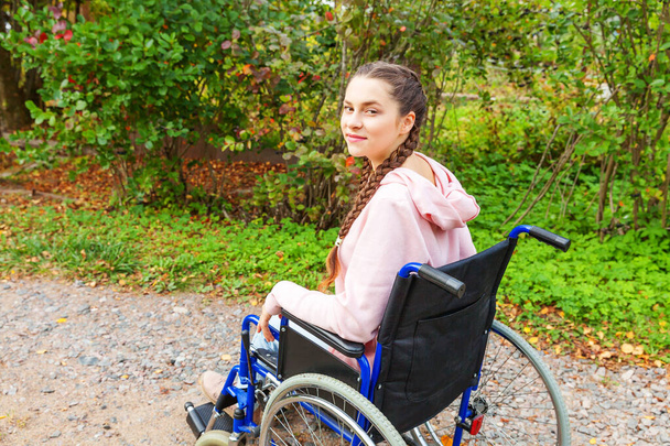 Young happy handicap woman in wheelchair on road in hospital park waiting for patient services. Paralyzed girl in invalid chair for disabled people outdoor in nature. Rehabilitation concept - Photo, Image