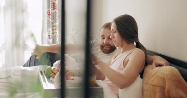 Happy couple lying on bed,laughing and watching funny photos on smartphone. Good looking man and woman having breakfast, and talking while looking at screen in bedroom. - Video, Çekim