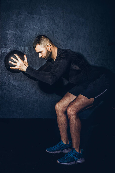 Man working out with his medicine ball on grey background. Bearded handsome young muscular sportsman is working out with a medicine ball in gym. High resulotion image. - Photo, Image