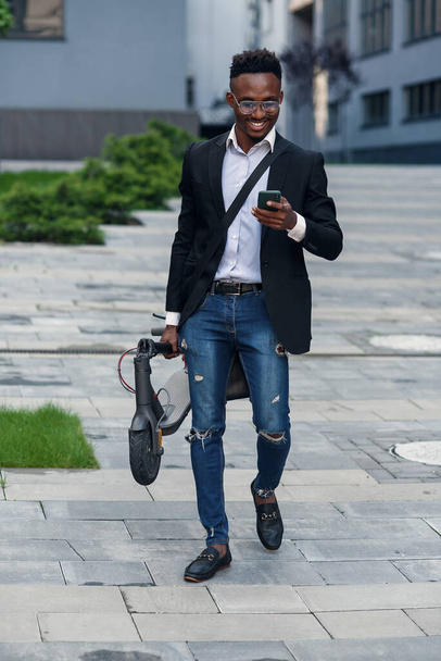 Empresário com scooter elétrico andando na frente do edifício de negócios moderno olhando para o telefone
. - Foto, Imagem