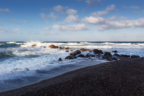 Santorini, Vlichada partja, a viharos tenger a part menti szikláknak ütközik. Magas hullámok, fehér hab és fekete homok a parton, gyönyörű kék ég fehér bárányokkal. - Fotó, kép