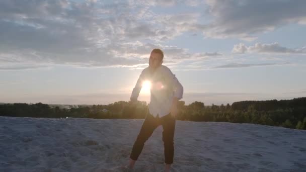 przystojny młody facet tańczący nowoczesny balet na piasku o zachodzie słońca w piękny letni wieczór. Współczesna choreografia. Zwolnij trochę. Kamera na żywo porusza się - Materiał filmowy, wideo