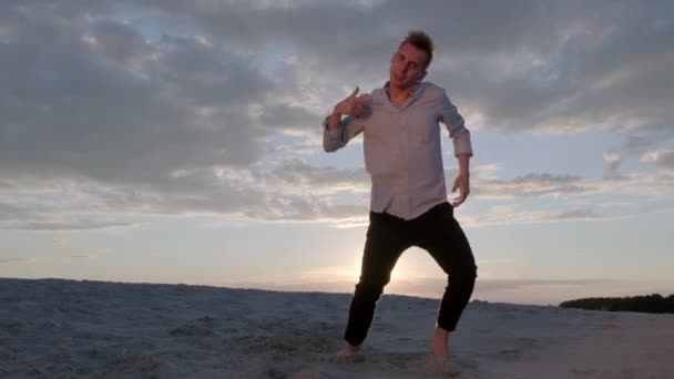 ハンサムなダンサーは日没時に砂浜でフリースタイルのダンス作曲を行います。コンテンポラリー振付。スローモーション映像。ライブカメラが動く - 映像、動画