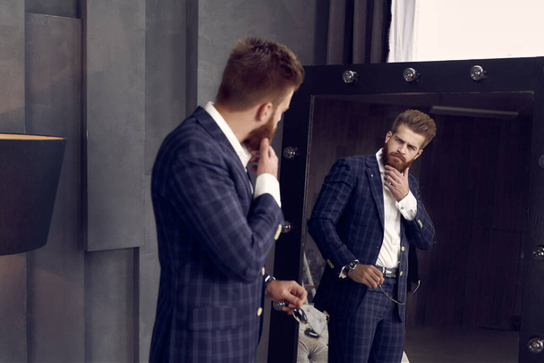 Schöner, stylischer Mann im blauen Anzug in einem Käfig zu Hause auf dem Sofa neben dem Spiegel stehend. Hält sein Kinn - Foto, Bild