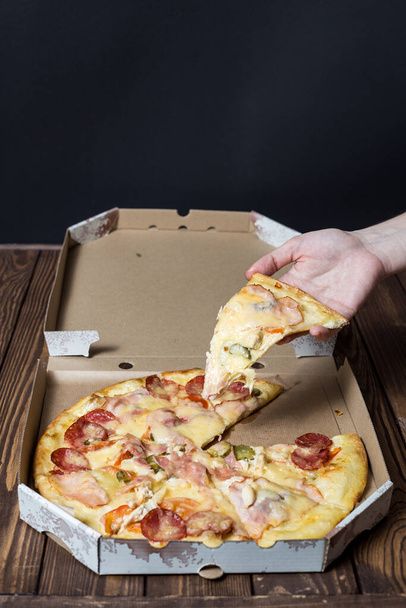 Το γυναικείο χέρι παίρνει ένα κομμάτι πίτσα. πίτσα σε ανοιχτό χαρτόκουτο σε ξύλινο τραπέζι. Φάστφουντ. διαφημιστική αφίσα. - Φωτογραφία, εικόνα