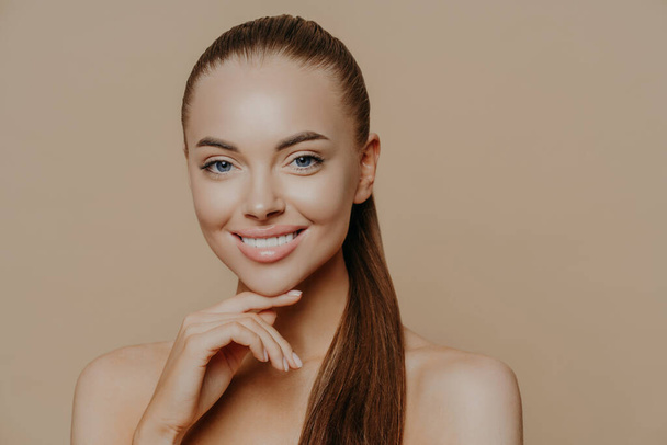 Το απομονωμένο πλάνο της νεαρής γυναίκας με ευρωπαϊκή εμφάνιση έχει καθαρό υγιές λείο δέρμα μετά από καθημερινές διαδικασίες καθαρισμού, χαμογελά με δόντια, στέκεται με γυμνούς ώμους σε εσωτερικό χώρο πάνω από το μπεζ τοίχο. - Φωτογραφία, εικόνα