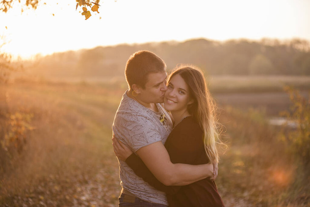 A young man kisses, hugs a beautiful smiling blonde girl with long hair. Close-up portrait of a couple embracing against the background of the autumn forest. Happy lovers in love travel together - Photo, image