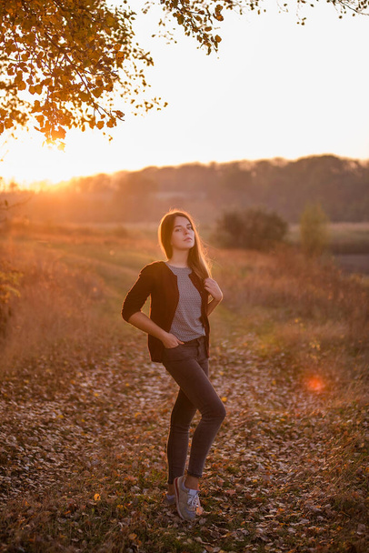 A modern, beautiful girl, in a burgundy jacket, dark jeans, stands in a field, blonde hair fluttering in the wind against the background of an autumn field at sunset. Autumn travel, rest from the city, freedom of thought. - Foto, imagen