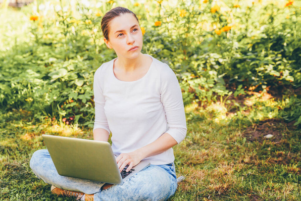 Мобильный офис. Концепция внештатного бизнеса. Молодая женщина, сидящая на лужайке в городском парке и работающая на ноутбуке. Аутентичная студентка по стилю жизни учится на открытом воздухе - Фото, изображение