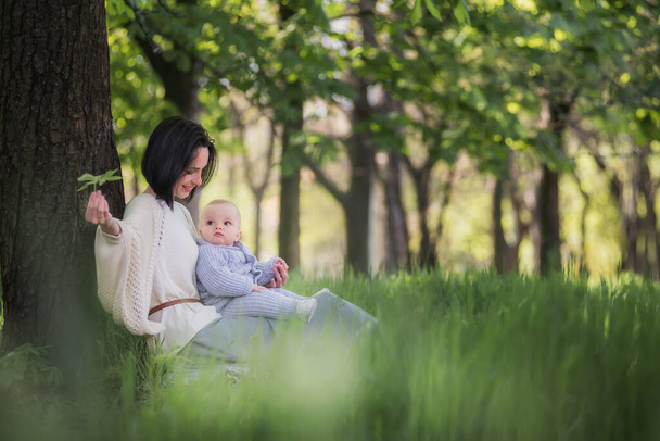 Modern beautiful young brunette mother with a short haircut, holding a toddler in a gray knitted romper in her arms. A girl and her son are sitting under a tree, playing together, hugging. Mom kisses the child - Foto, Bild