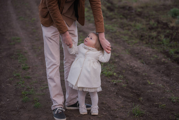 Отец и маленькая дочь ходят под руку в цветущих садах на закате. Молодой человек обводит девушку за руки, она смеется. Отношения между родителями и детьми, воспитание. Счастливая семья - Фото, изображение