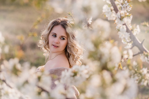 Delicate, dreamy girl with curly blond hair in blooming white gardens at sunset. Close-up portrait with nude makeup, clean face of a young woman in a light blue dress. Walk, rest outside the city - Фото, изображение