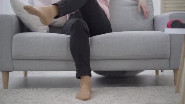Unrecognizable woman sitting on couch and crossing legs. Confident adult Caucasian lady in black pants resting indoors. Lifestyle, relaxation, leisure. - Filmati, video