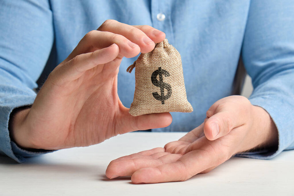 Uma mão em uma camisa segura um pequeno saco com dinheiro e um sinal de dólar dos EUA
 - Foto, Imagem