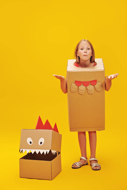 Emotional little girl playing with a cardboard dragon, dinosaur. Childhood dreams. Fantasy, imagination. Studio portrait on a yellow background. - Fotó, kép
