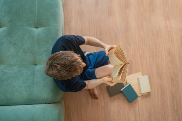 κορυφαία άποψη του νεαρού άνδρα που κάθεται στο ξύλινο πάτωμα και διαβάζοντας το βιβλίο στο σπίτι - Φωτογραφία, εικόνα