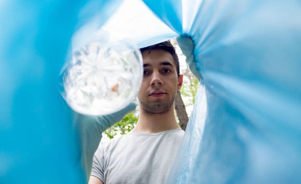 Ein junger Mann aus dem Kaukasus steckt eine Plastikflasche in einen Müllsack. Der Blick aus der Tasche. Müllabfuhr im Wald. - Foto, Bild