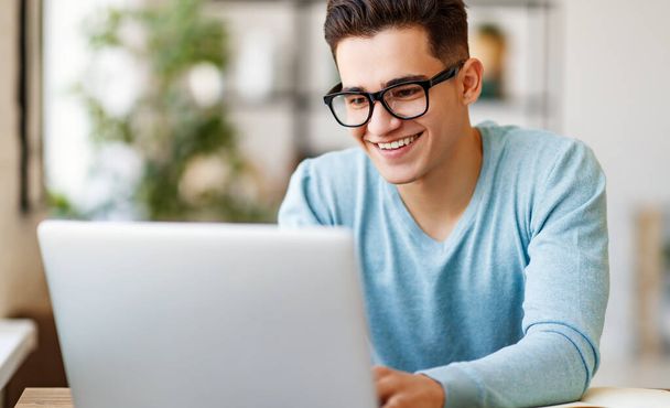 Šťastný etnický mladý muž s úsměvem a prohlížení dat na notebooku, zatímco dělá domácí projekt u stolu u hom - Fotografie, Obrázek