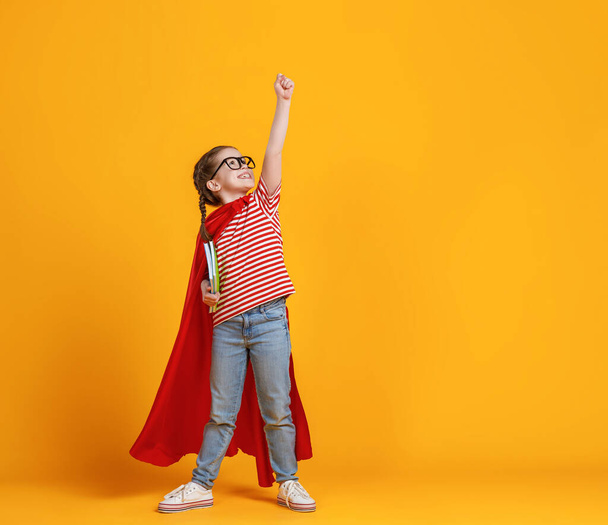 Koko kehon tyttö supersankari viitta hymyillen ja nostamalla nyrkki ylös ollessaan valmis koulun tutkimukset vastaan keltainen backdro - Valokuva, kuva