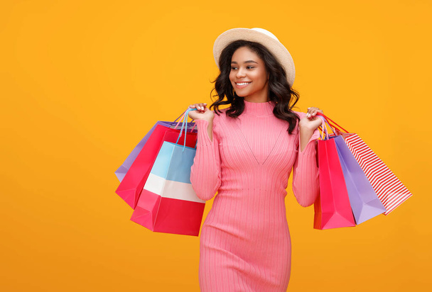 Side view of cheerful ethnic woman with paper bags smiling and looking away over shoulder while walking against yellow backdrop after shoppin - Photo, Image