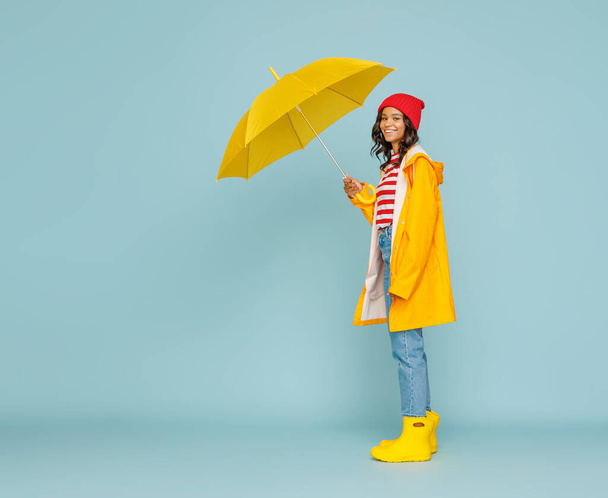 Positive ethnische Frau mit Hut und Regenmantel lächelt und blickt in die Kamera, während sie an einem regnerischen Tag vor rotem Hintergrund unter einem Regenschirm steht - Foto, Bild