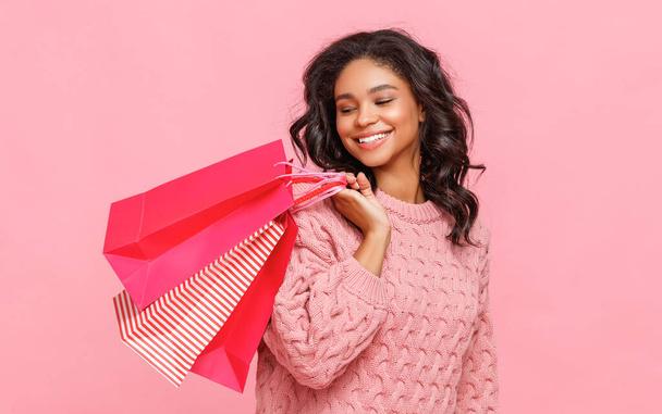 Mujer étnica positiva sonriendo para la cámara y llevando bolsas de papel en el hombro durante las compras contra el fondo rosa - Foto, imagen