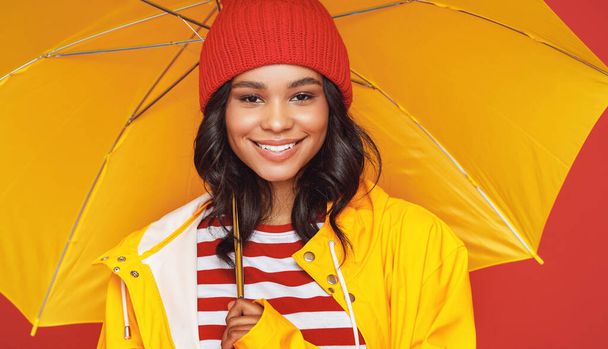 Femme ethnique positive en chapeau et imperméable souriant et regardant la caméra tout en se tenant sous le parapluie le jour de pluie contre fond rouge - Photo, image