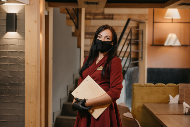 A female restaurant manager in black disposable medical gloves wears a medical face mask is posing holding a wooden menu in a restaurant. A cafe owner with long hair in a ruby dress. - Photo, Image