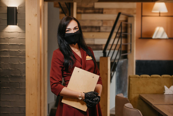 A female restaurant manager in black disposable medical gloves wears a medical face mask is posing holding a wooden menu in a restaurant. A charming cafe owner with long hair in a ruby dress. - Photo, Image