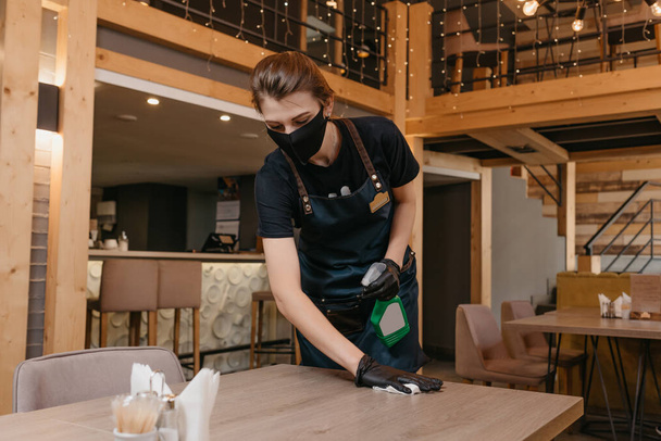 A waitress who wears a black medical face mask and disposable medical gloves is holding a bottle with sanitizer and cleaning a table with a rag in a restaurant. A barista is waiting for clients. - Photo, Image