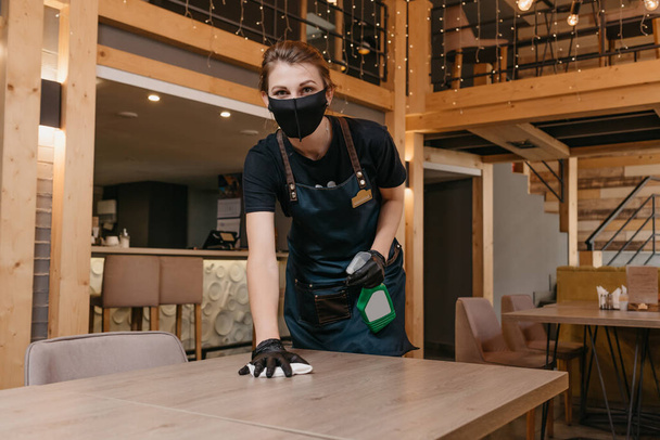 A kind waitress who wears a black medical face mask and disposable medical gloves is holding a bottle with sanitizer and cleaning tables with a rag in a restaurant. The staff is waiting for clients. - Photo, Image