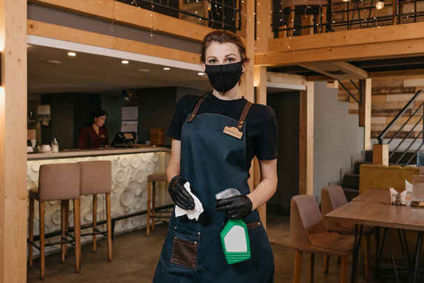 Een stijlvolle serveerster die een schort, een zwart medisch gezichtsmasker en wegwerphandschoenen draagt, houdt een fles met ontsmettingsmiddel en een doek vast in een restaurant. Het personeel wacht op klanten in een café. - Foto, afbeelding
