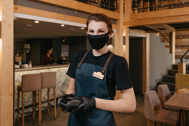 A waitress who wears an apron, a black medical face mask, and disposable medical gloves is holding a wireless payment terminal in a restaurant. The staff is waiting for the client to pay a bill. - Photo, Image