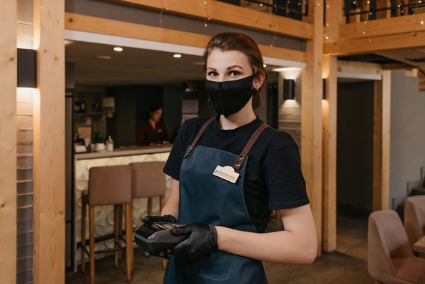 Μια χαριτωμένη σερβιτόρα που φοράει ποδιά, μαύρη μάσκα προσώπου και γάντια μιας χρήσης κρατάει ένα ασύρματο τερματικό πληρωμής σε ένα εστιατόριο. Το προσωπικό περιμένει τον πελάτη να πληρώσει ένα λογαριασμό.. - Φωτογραφία, εικόνα