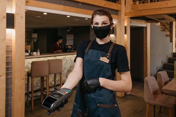 A waitress who wears an apron, a medical face mask, and disposable medical gloves is handing a wireless payment terminal to a client in a restaurant. The staff is waiting for customer to pay a bill. - Photo, Image