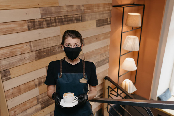 Een mooie serveerster die een zwart gezichtsmasker draagt en wegwerphandschoenen houdt een kop koffie vast in een restaurant. Een schattige barista wacht op klanten in een café. - Foto, afbeelding