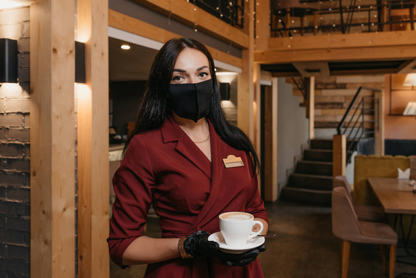 Розкішна жінка-менеджер ресторану, яка носить чорну маску для обличчя і одноразові рукавички, тримає чашку кави в ресторані. Красивий власник кафе в рубіновому платті чекає клієнтів
. - Фото, зображення