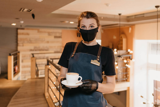 Una cameriera che indossa una maschera nera e guanti monouso sta servendo una tazza di caffè al secondo piano di un ristorante. Un barista carino è in attesa di clienti in un caffè. - Foto, immagini