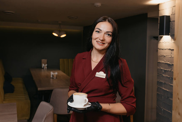 Egy gyönyörű női étterem menedzser fekete, eldobható kesztyűben egy csésze kávét szolgál fel egy kávézóban. Egy rubin ruhás kedves kávézó várja az ügyfeleket.. - Fotó, kép