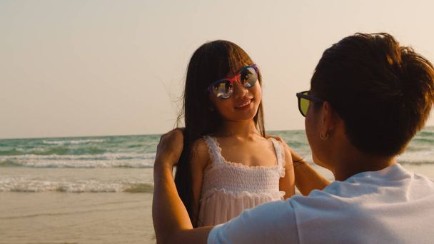 アジアの若い幸せな家族は夕方にビーチで休暇を楽しむ。お父さんは日没時に一緒に海の近くでリラックスしながらサングラスをかけるのを助けます。ライフスタイル旅行休暇夏旅行のコンセプト. - 写真・画像