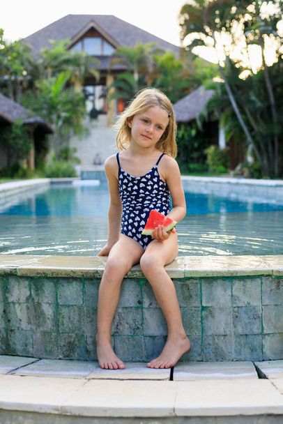 Roztomilá holčička v modrých plavkách sedící u bazénu a držící kousek melounu. Užij si to tropické ovoce. Letní koncept. Šťastné dětství. Trávit čas venku. Bali, Indonésie - Fotografie, Obrázek
