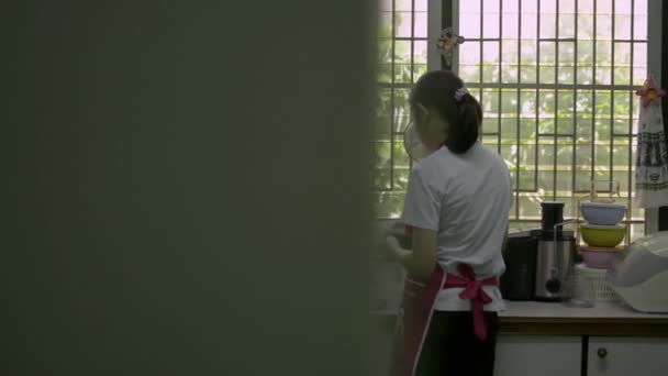 Chica asiática con delantal ayudando a su madre a preparar y limpiar los utensilios en el armario de la cocina al lado de la ventana por la mañana. Trabajo doméstico. Panorámica tiro
. - Metraje, vídeo