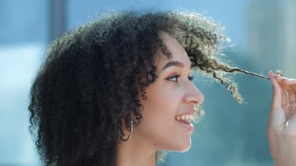 Close-up face african girl curly hairstyle. Young American playful woman holding curls hair in hands, toothy smile. Coiffed hair brushing styling dry brittle hair in afro braids, hair frizz care - Materiał filmowy, wideo