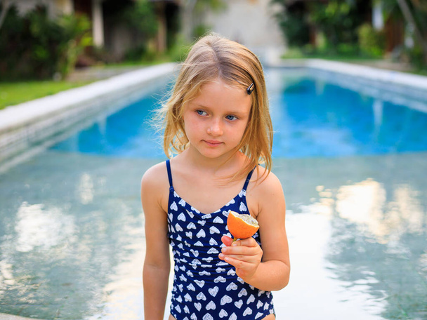 Roztomilé blonďaté děvčátko v modrých plavkách sedící u bazénu a držící v náručí vášnivé ovoce. Užij si to tropické ovoce. Letní koncept. Šťastné dětství. Dovolená v Asii. Bali, Indonésie - Fotografie, Obrázek