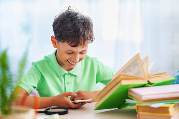 lazy kid enthusiastically plays game on smartphone instead homework, discipline. boring little schoolboy is playing with his smartphone instead doing homework. Concept of Internet and gaming addiction - Photo, image